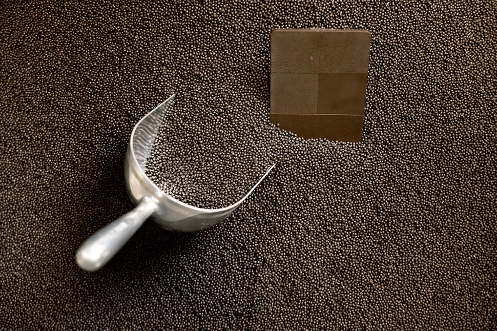 A metal scoop rests on a large pile of small, round pellets, with a square section of flat metal embedded in the pellets nearby.