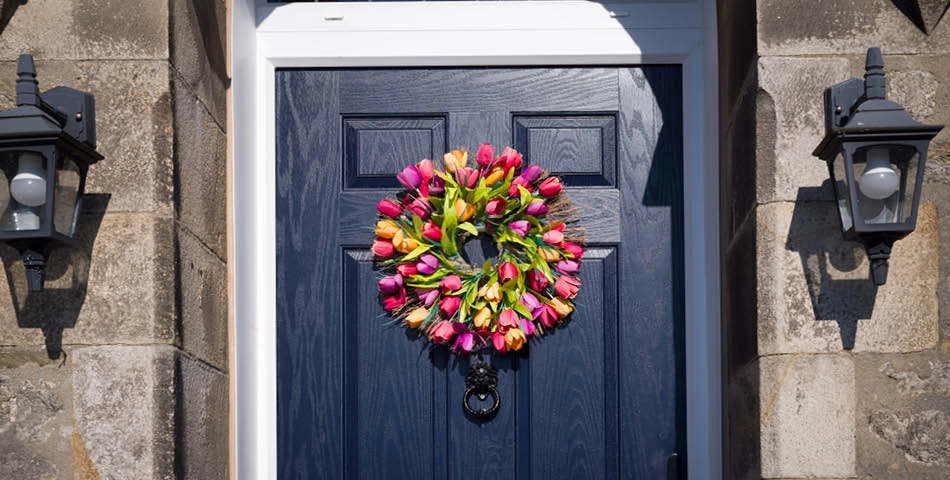 A dark blue front door with a colorful wreath made of pink, orange, and yellow tulips, flanked by two black wall lanterns on a stone exterior.