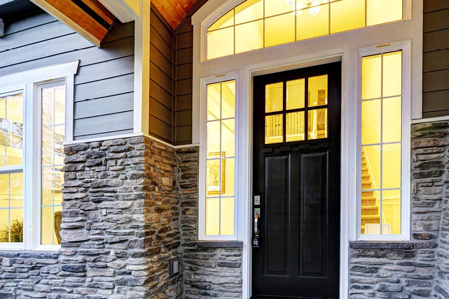 Front entrance of a house with a black door, large windows, stone veneer siding, and warm interior lighting visible through the glass.