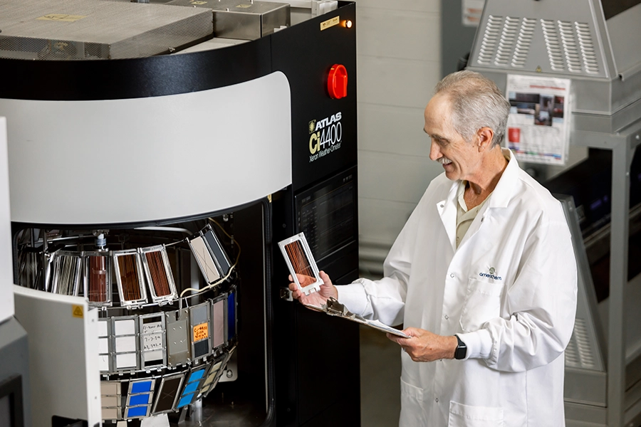A scientist in a lab coat examines test samples in front of a large testing machine, holding a clipboard and looking at a sample panel.
