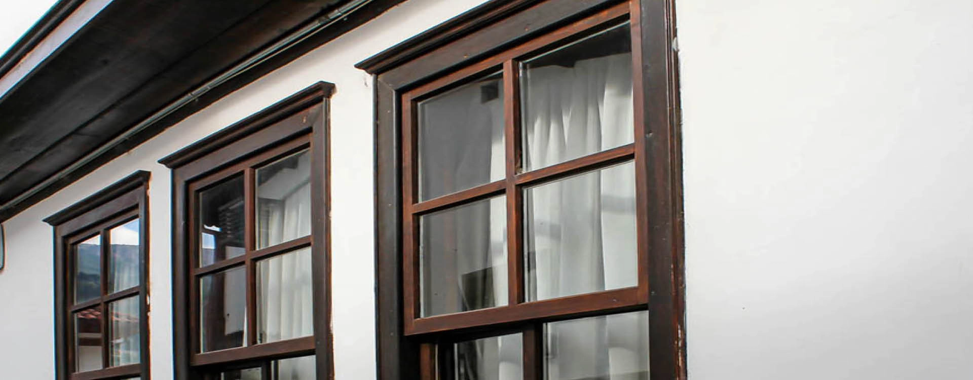 Three wooden-framed windows with white curtains are set in a white exterior wall of a building.