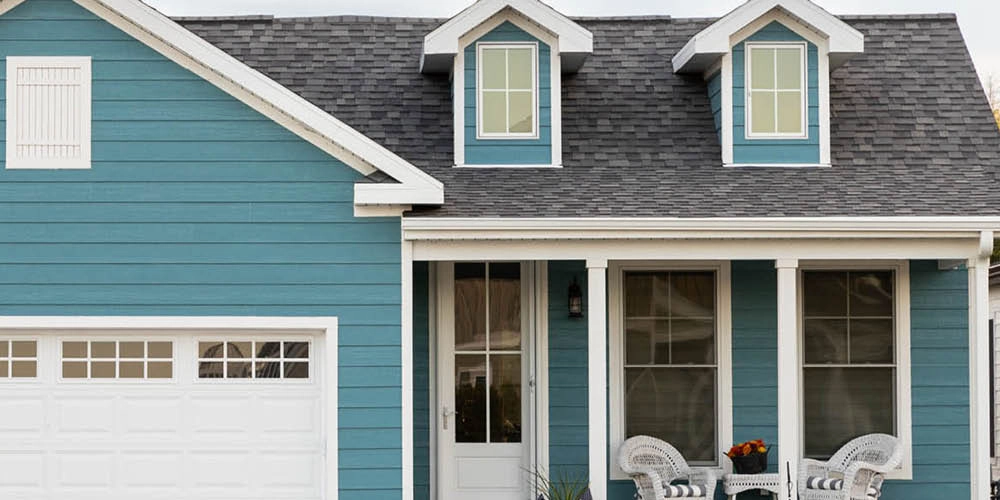 Blue house exterior with white trim, two upper dormer windows, a white garage door, and a small porch with two white chairs and a table.