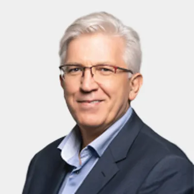 A man with short gray hair and glasses, wearing a dark suit jacket over a light blue shirt, smiles at the camera against a plain light background.