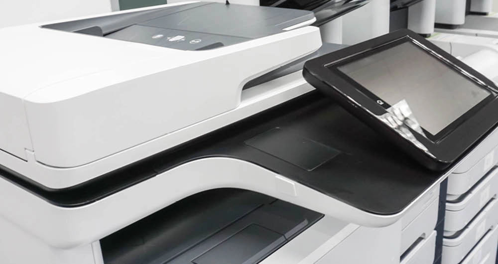 A close-up view of a modern office printer or copier with a touchscreen control panel and paper trays.