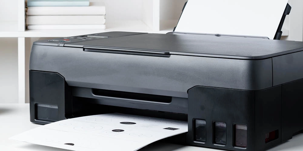 A black inkjet printer prints a sheet of paper in an office setting with books on a white shelf in the background.