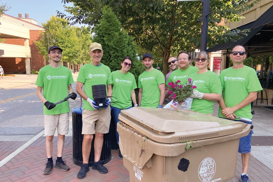 A group of seven people in green shirts stand on a sidewalk near a brown compost bin, holding gardening tools and flowers, with trees and buildings in the background.
