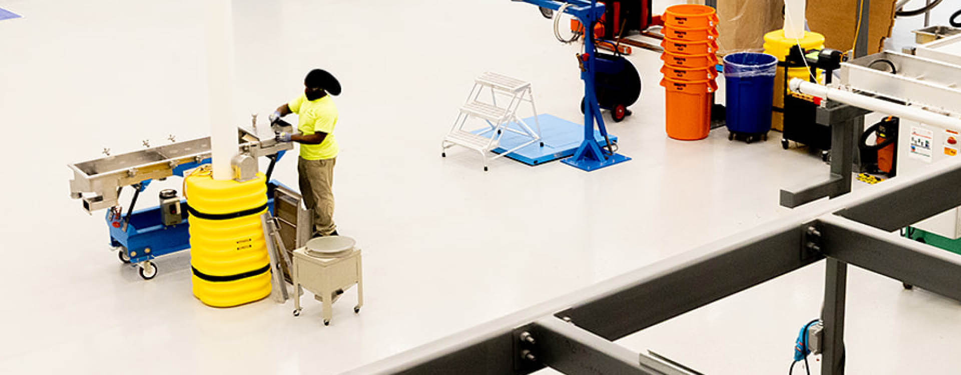 A worker in safety gear operates machinery next to yellow barrels and various industrial equipment in a clean, spacious facility.