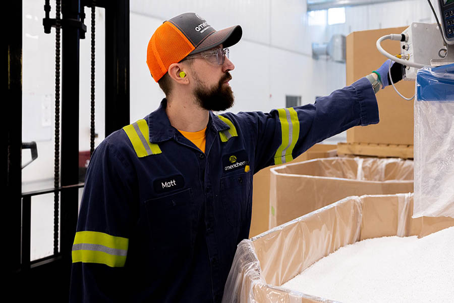 A worker in safety gear operates machinery in an industrial setting, standing next to a large container filled with white granular material.