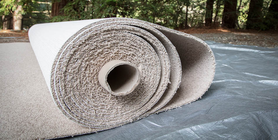 A large beige carpet roll lies partially unrolled on a tarp outdoors, with trees visible in the background.