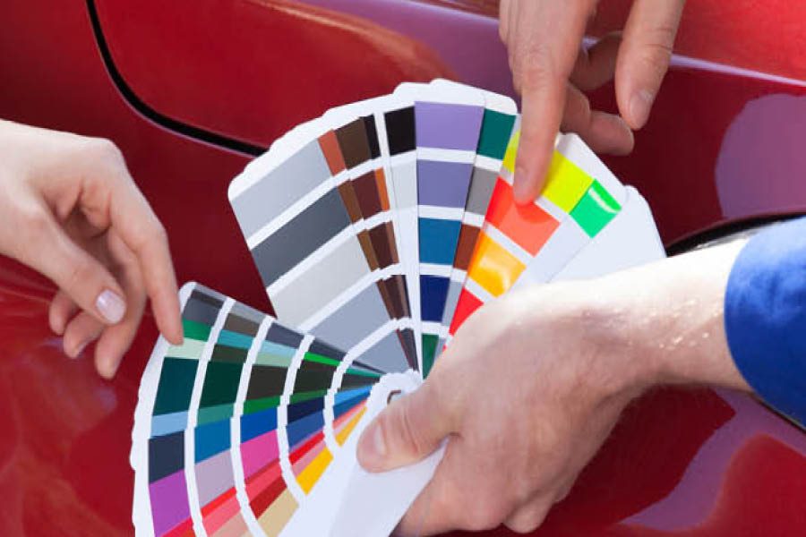 Three people hold and compare color swatches against the red surface of a vehicle, possibly selecting a paint color.