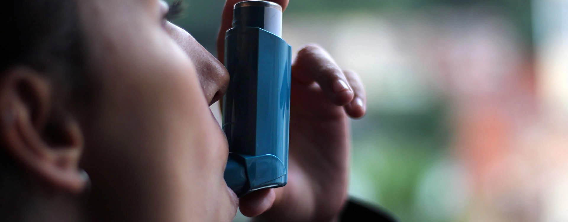 A person uses a blue inhaler, holding it to their mouth and pressing down to release medication.