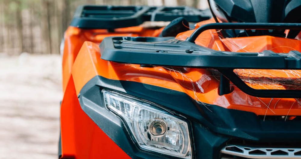 Close-up of the front left side of an orange all-terrain vehicle (ATV) with visible headlight, grill, and black trim.