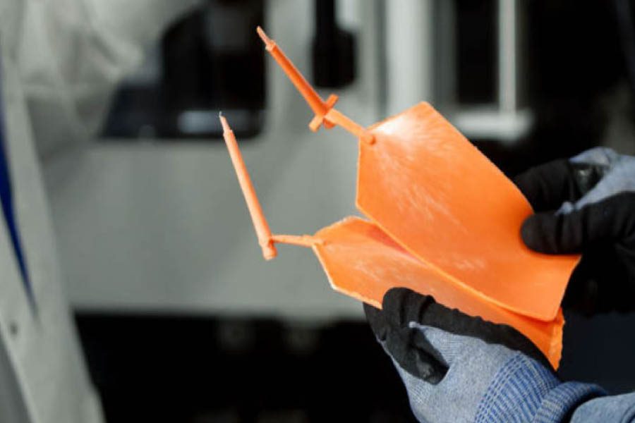 Gloved hands hold two orange plastic injection mold parts with attached runners and sprues, set against a blurred industrial background.