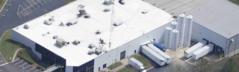 Aerial view of a large industrial building with a white roof, multiple loading docks, silos, and several trucks parked outside.
