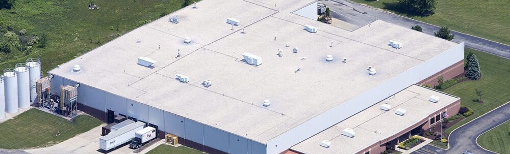 Aerial view of a large industrial warehouse with a flat white roof, loading dock area, parked trucks, and storage tanks on a grassy lot.