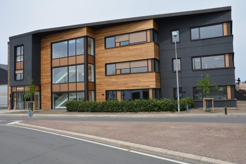 A modern three-story building with large windows, wood paneling, and black accents, situated on a corner next to a road with minimal landscaping.