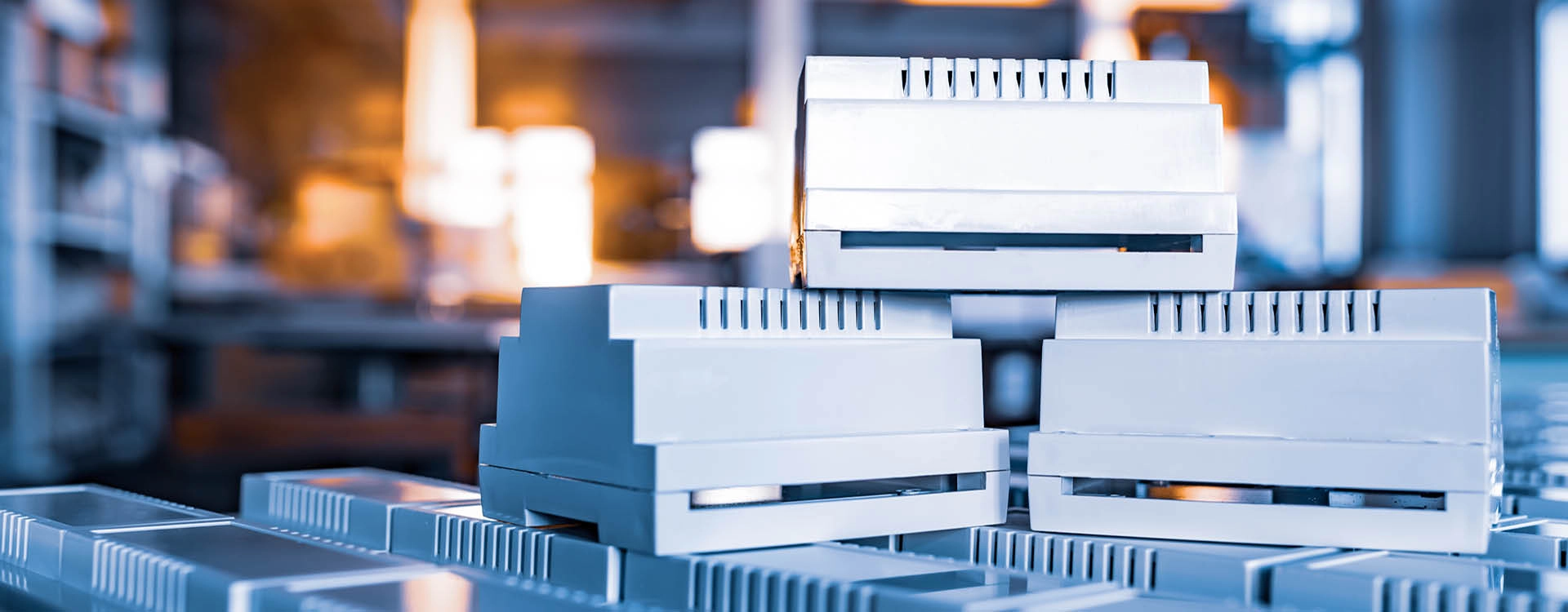 Stacked gray electronic DIN rail enclosures in a factory or industrial setting, with blurred background and warm lighting.