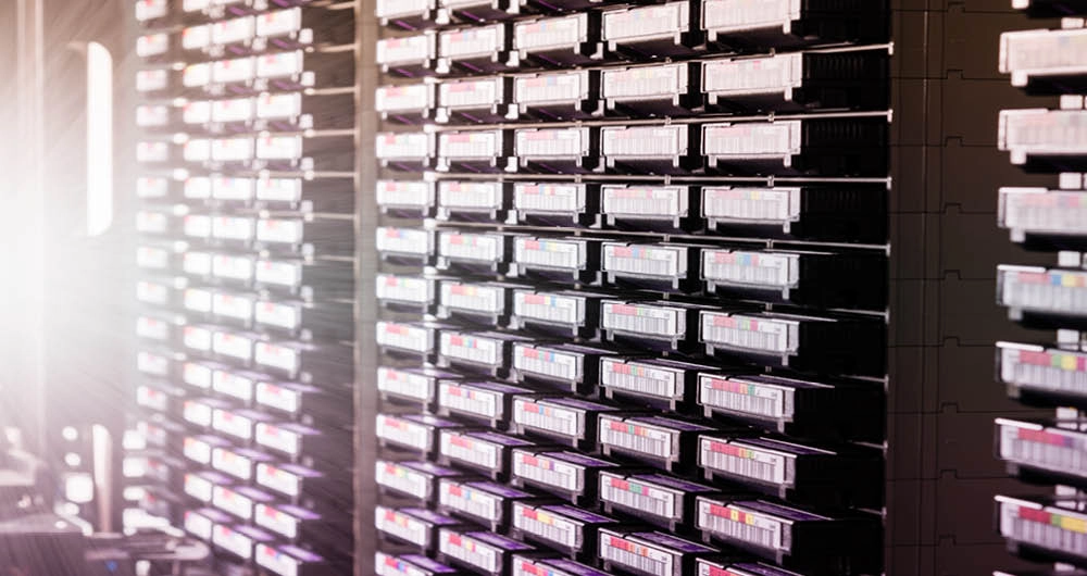 Server racks filled with multiple rows of networked hardware equipment are shown in a brightly lit data center.