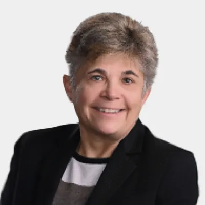 Person with short, gray hair wearing a black blazer and white shirt, smiling at the camera against a plain, light background.