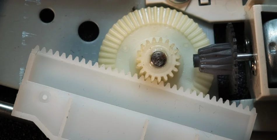 Close-up of interlocking plastic gears in a mechanical device, showing a white rack and pinion system with a gray gear and metal components.