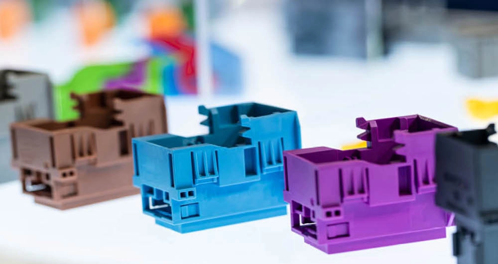 Three plastic components in brown, blue, and purple are arranged in a row on a white surface, with a blurred colorful background.