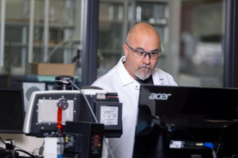 A man in a lab coat and safety glasses works at a computer station in a laboratory setting with equipment visible around him.