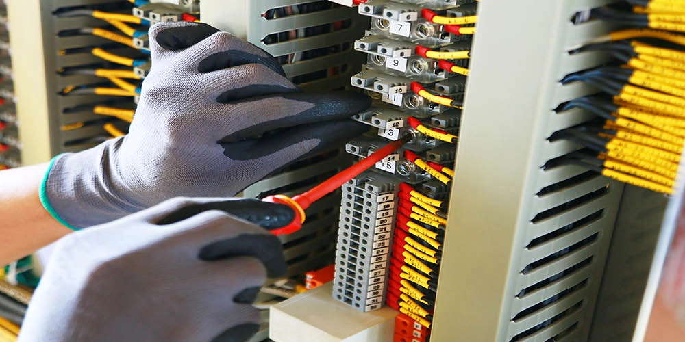 A person wearing gloves uses a screwdriver to work on wiring connections inside an electrical control panel.