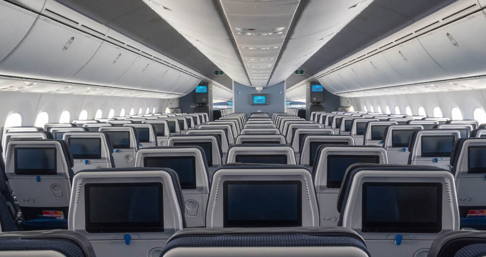 Rows of empty airplane seats with individual screens, seen from the back of the cabin, facing towards the front of the aircraft.