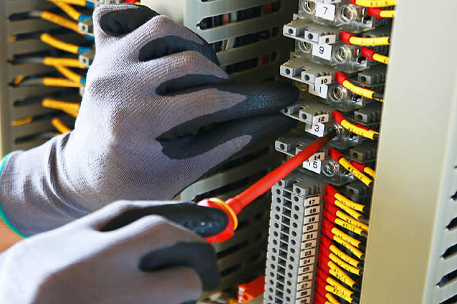 A person wearing gloves uses a screwdriver to adjust wires and terminals in an electrical control panel.