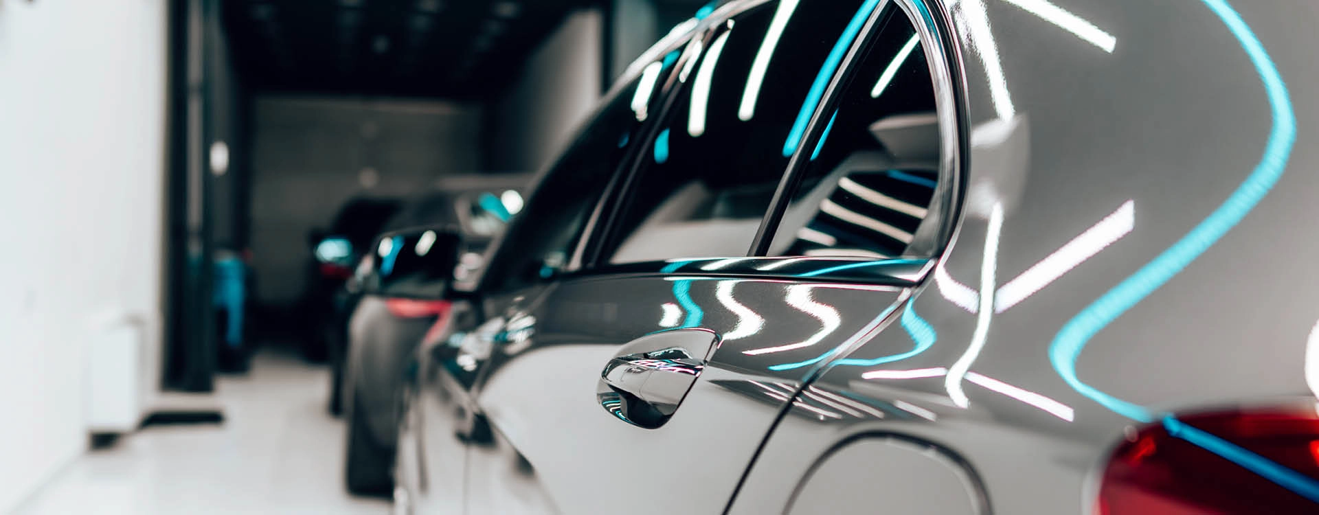 Close-up side view of a shiny, silver car with reflections of overhead lights, parked indoors alongside other vehicles.
