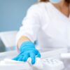 Person wearing blue gloves and a mask cleans a white surface with a disposable wipe in a clinical or medical setting.