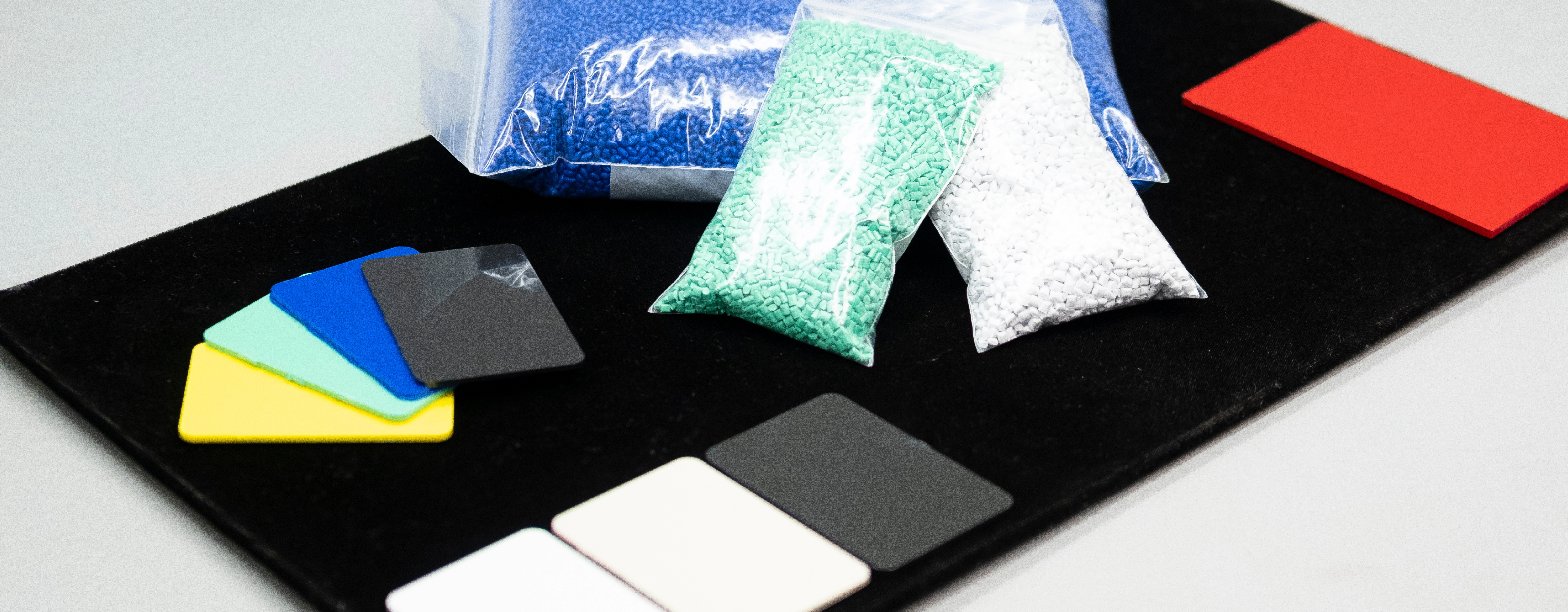 Bags of colored plastic pellets and several rectangular plastic samples in various colors arranged on a black surface.