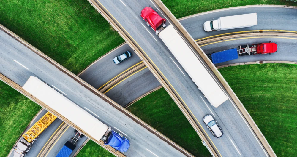 Aerial view of intersecting highways with several semi-trucks and cars driving on multiple lanes over green grass.