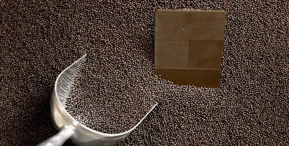 A metal scoop rests on a pile of small, round pellets with a square metal object partially uncovered among them.
