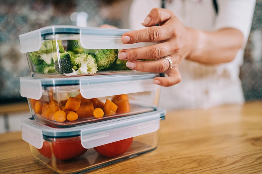 A person stacks three food containers with vegetables: broccoli, carrots, and tomatoes on a wooden countertop.
