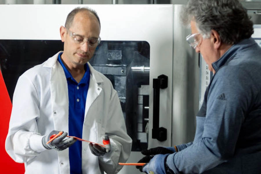 Two men wearing safety glasses and gloves inspect a long orange plastic piece in an industrial setting with machinery in the background.