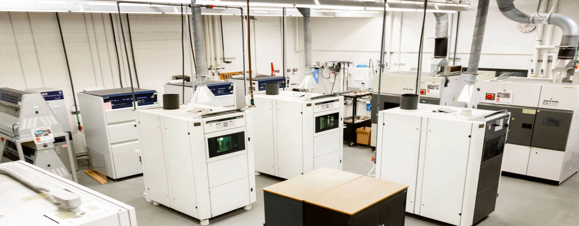 A laboratory room with several large industrial 3D printers and related equipment, arranged in rows under bright overhead lighting.