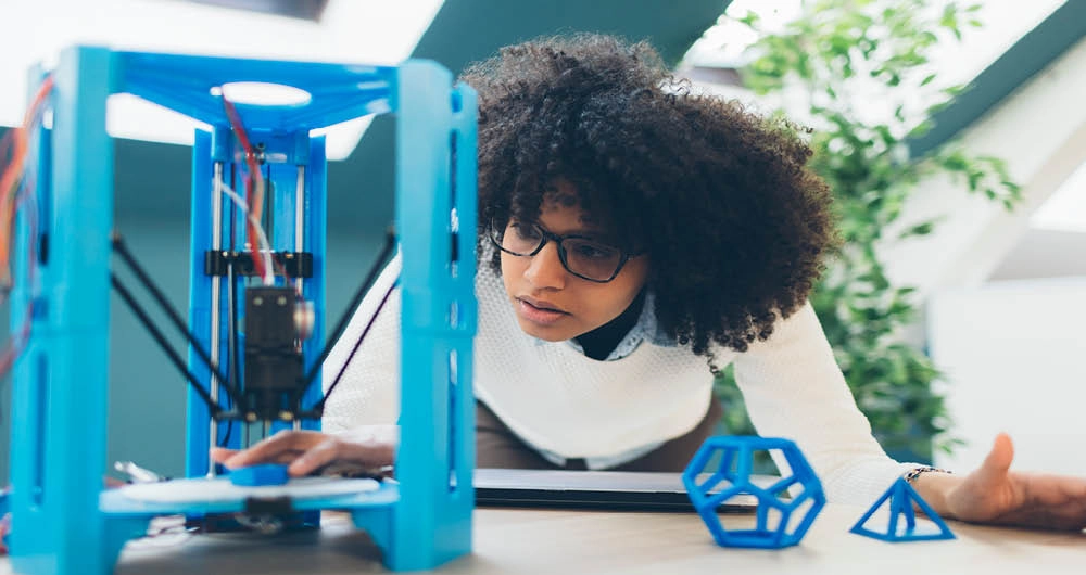 A person examines a blue 3D printer in operation, with two geometric models placed on the table nearby.