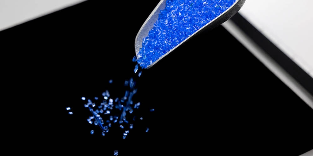 A close-up of blue granules being poured from a metal scoop onto a black surface.