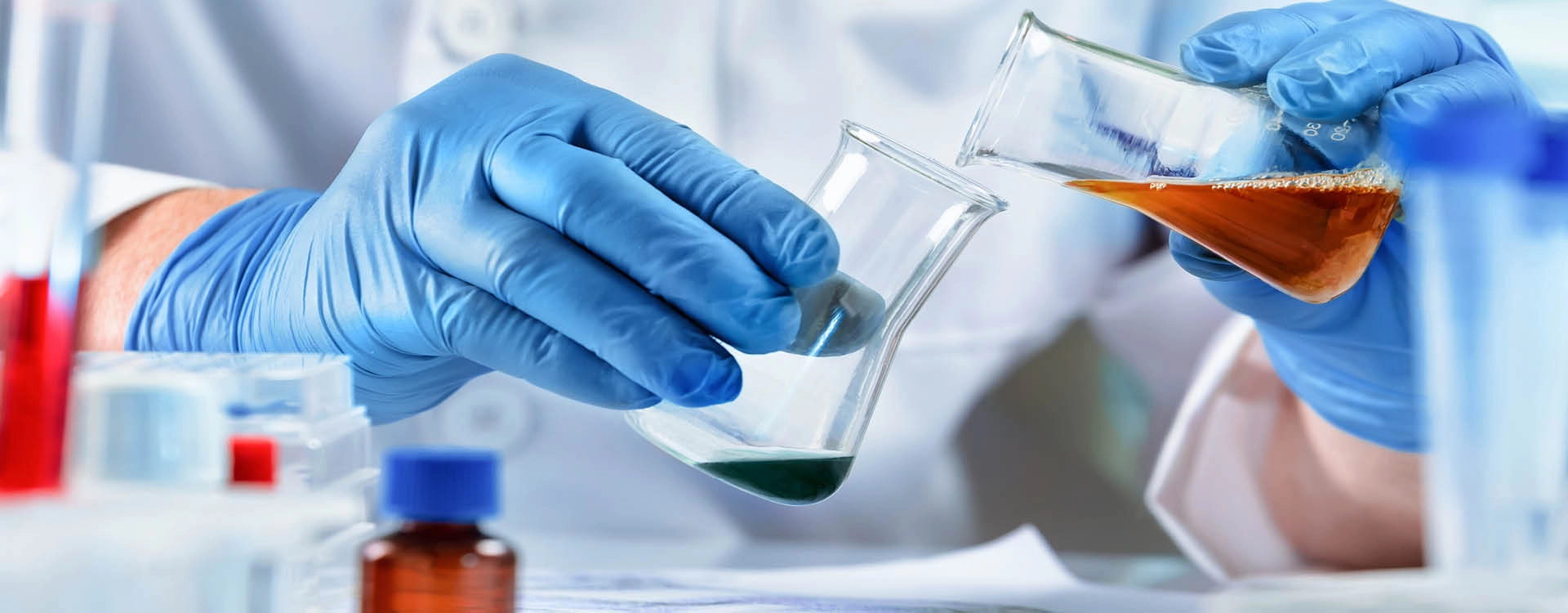 A scientist wearing blue gloves pours a liquid from a beaker into another container in a laboratory setting.