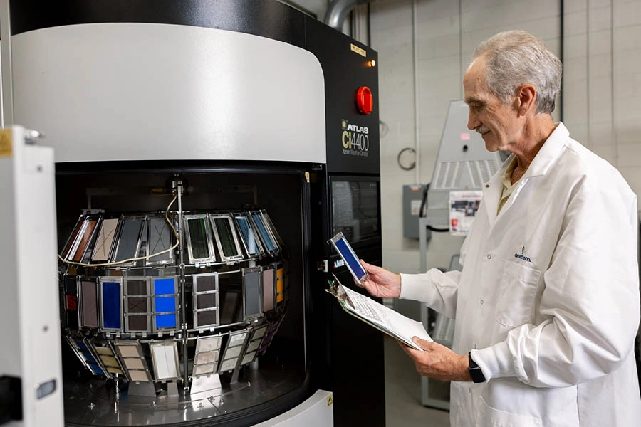 A man in a lab coat inspects a rotating machine holding color samples, taking notes on a clipboard in an industrial laboratory setting.