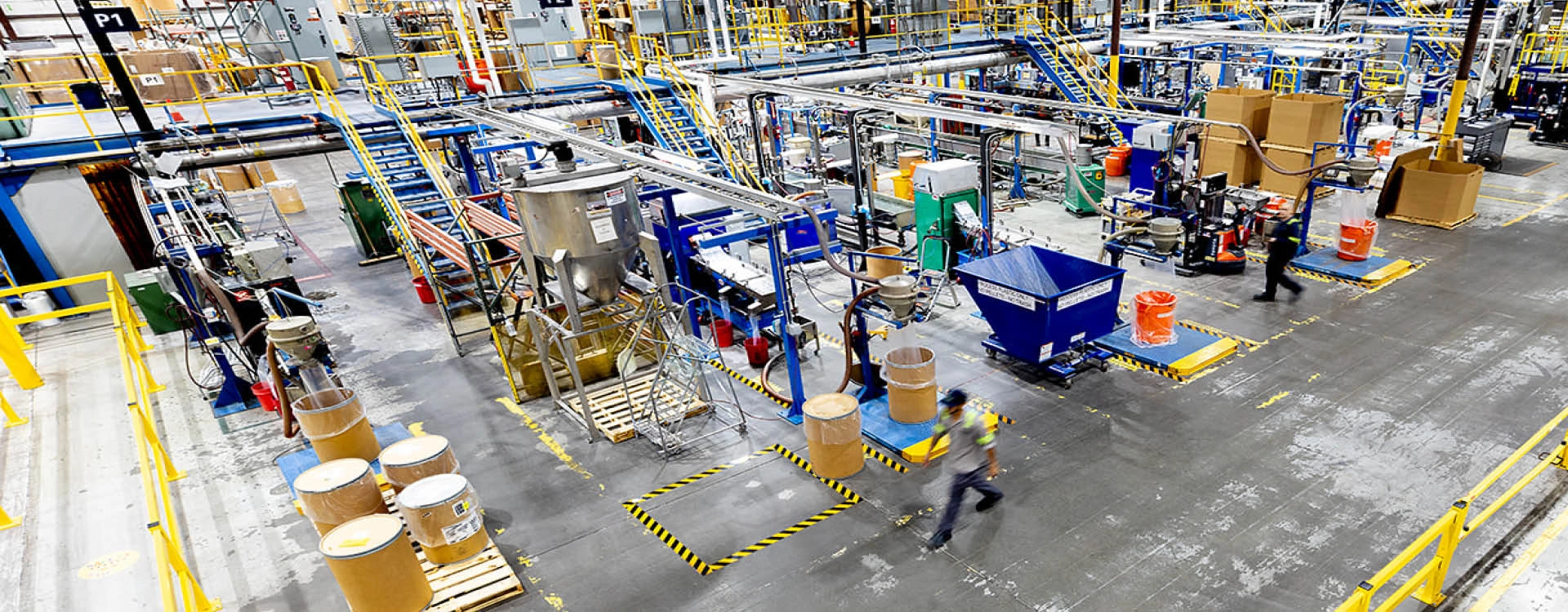 Large industrial factory interior with machinery, conveyor belts, cardboard barrels, and workers wearing safety gear moving through the space.