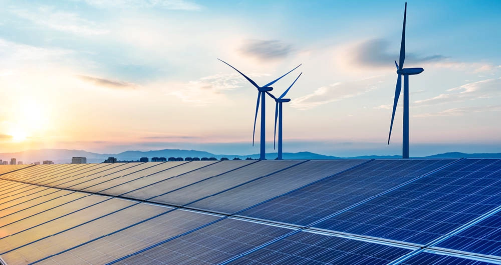 Rows of solar panels in the foreground with three wind turbines in the background under a clear sky at sunrise or sunset.