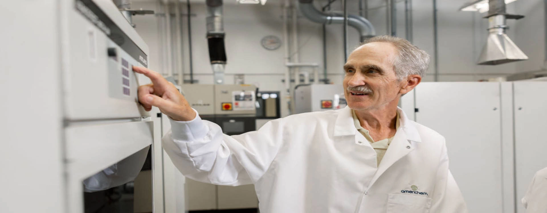 A man in a white lab coat operates a control panel in a laboratory with industrial equipment visible in the background.