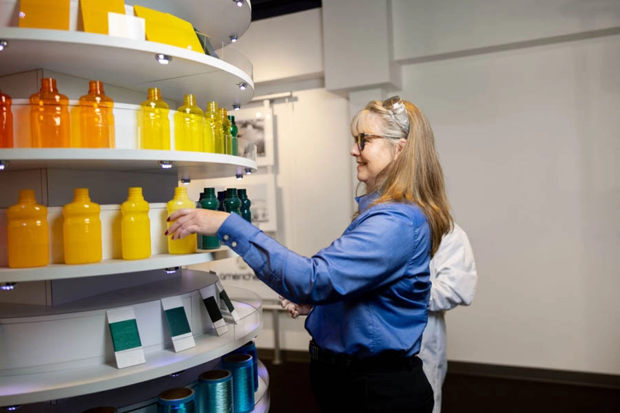 A woman in a blue shirt selects a yellow bottle from a display shelf arranged with bottles and swatches in gradient colors.