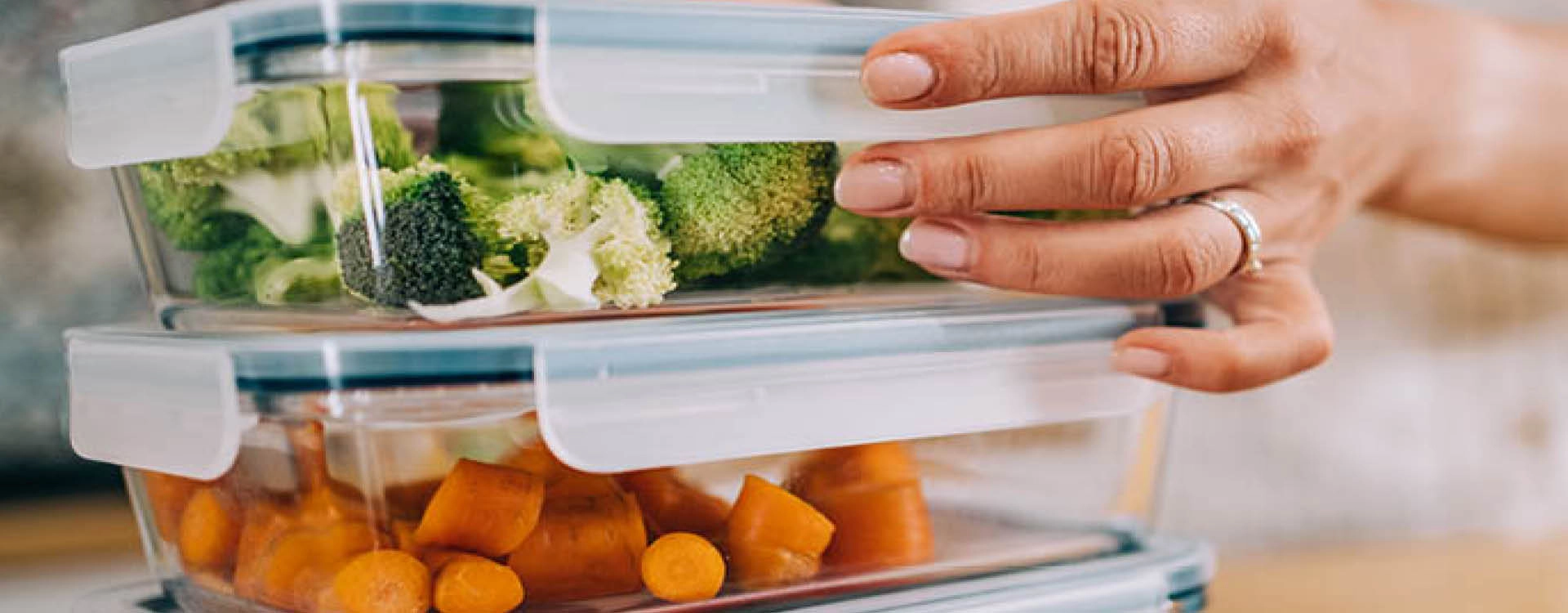 A hand holds a stack of glass food containers filled with chopped broccoli and carrots, with airtight lids.