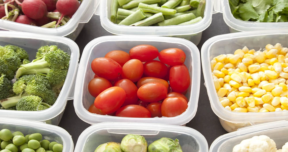 Assorted fresh vegetables, including cherry tomatoes, green beans, corn, broccoli, and radishes, packed in clear plastic containers.