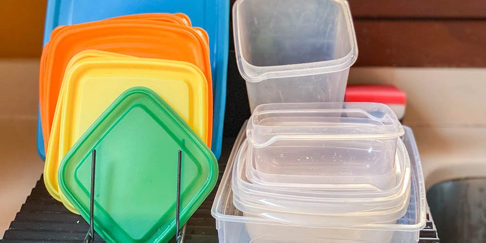 Plastic food storage containers and lids in various colors and sizes are drying on a dish rack next to a kitchen sink.