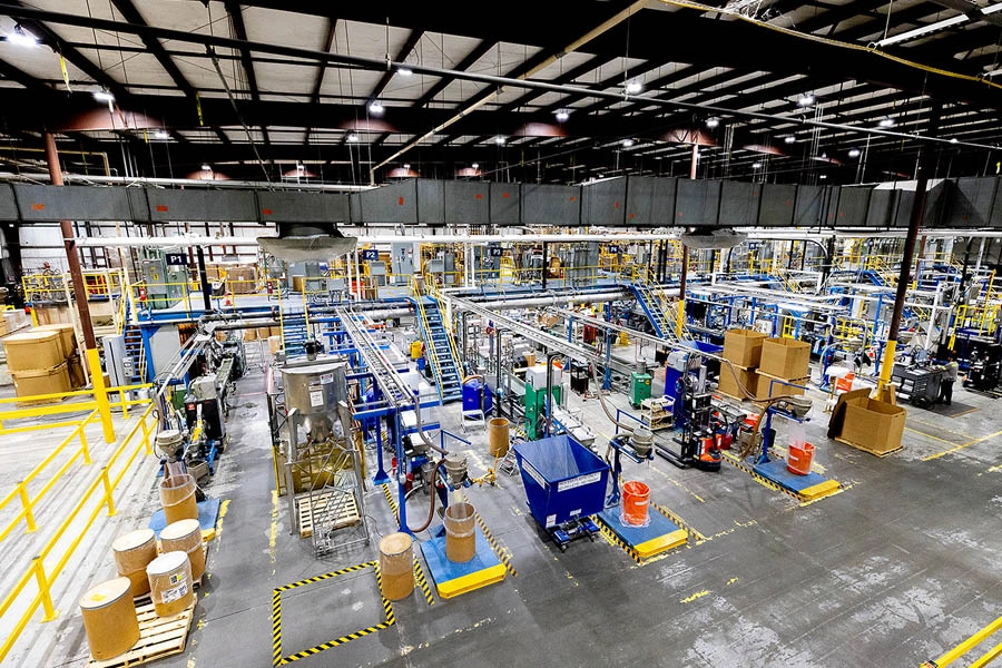 Large industrial factory interior with machines, conveyor belts, cardboard drums, and stacked pallets under bright overhead lighting.