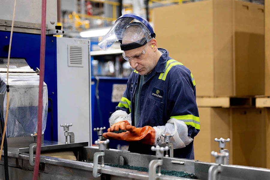 A worker wearing safety gear operates machinery in an industrial facility, using gloved hands and standing next to large equipment and cardboard boxes.